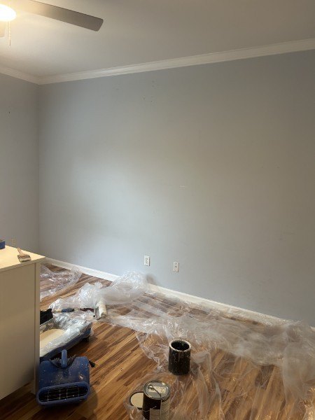 Light gray bedroom wall with white baseboard, painting supplies on the wood floor